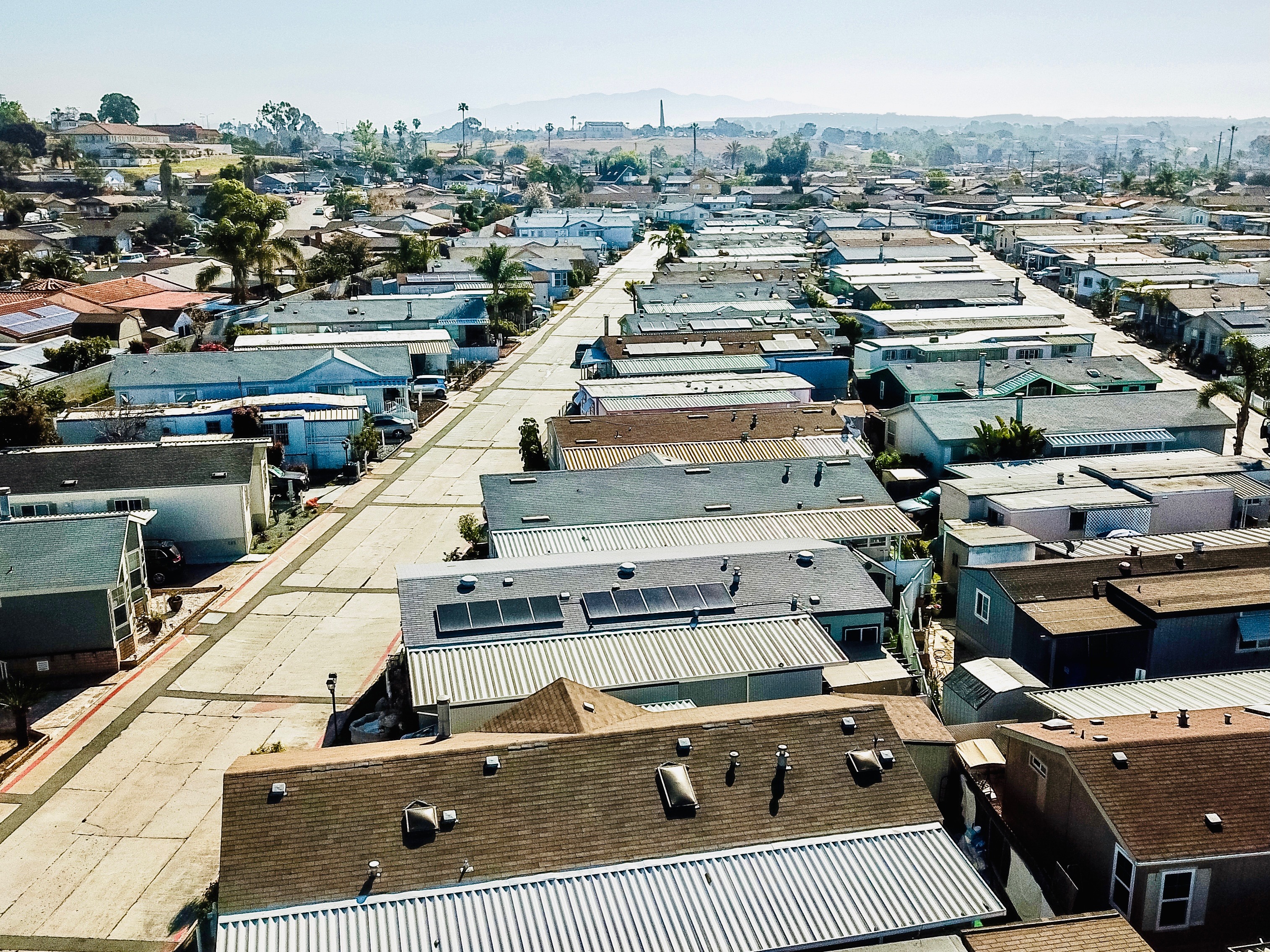 Aerial view of a mobile home community in Southern California, showcasing multiple manufactured homes with solar panel installations on rooftops, surrounded by palm trees and residential streets.